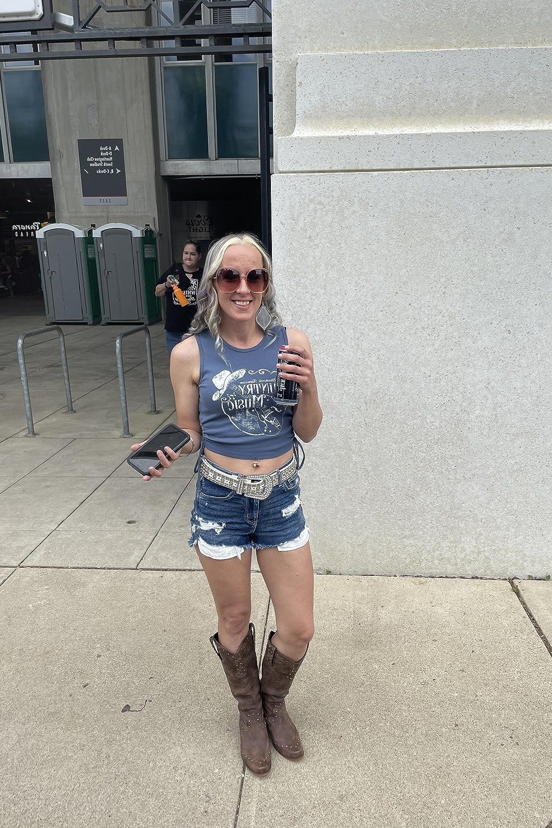 smiling blonde woman in her 40s wearing a cropped blue graphic tank, distressed denim shorts, a wide concho belt and brown cowboy boots outside a venue