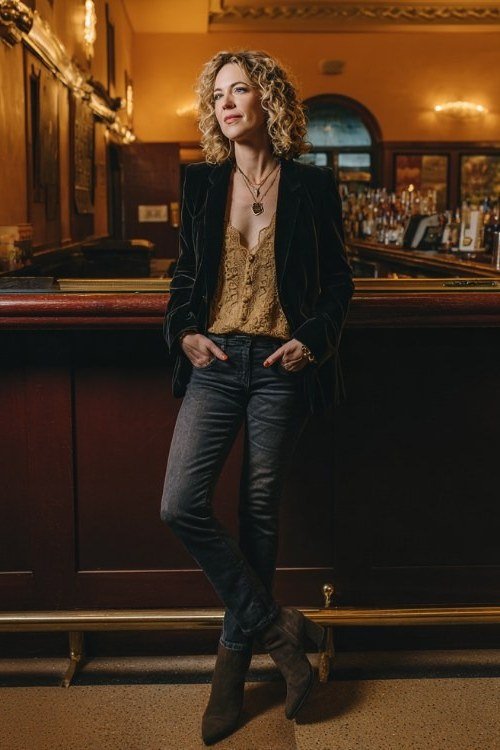 curly blonde woman in her 40s leaning against a bar in a black velvet blazer over a gold lace-trim top, dark skinny jeans and brown ankle boots