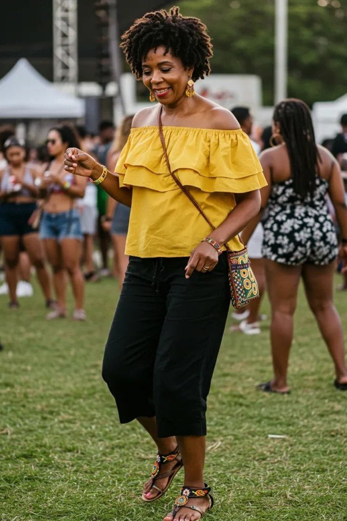 curly-haired woman dancing at a festival in a bright yellow off-shoulder ruffle top, black capri culottes and strappy flat sandals