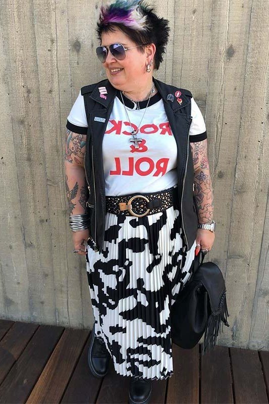 tattooed woman in her 40s with a colorful mohawk wearing a white rock and roll tee under a patched black vest, a cow-print pleated midi skirt, studded belt and black boots