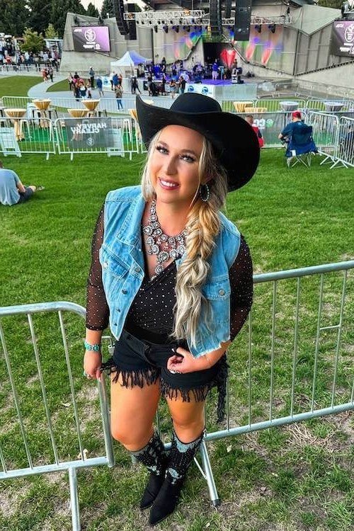 smiling blonde woman sitting on a festival rail in a black cowboy hat, a cropped denim vest with fringe over a black sequin top, black fringe shorts and black cowboy boots