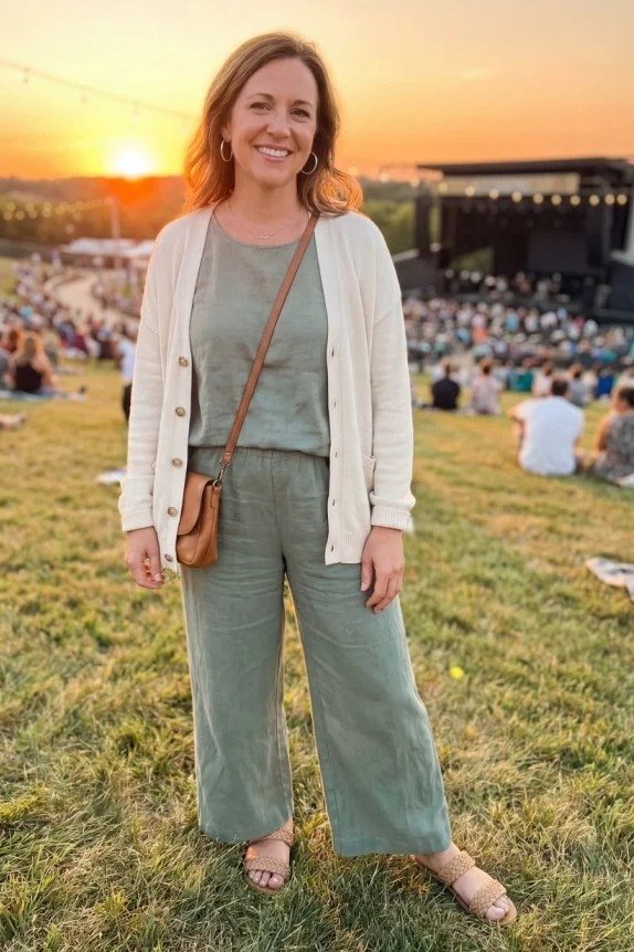 woman in her 40s at a sunset concert in a sage-green wide-leg jumpsuit layered with an open cream cardigan, a brown crossbody and flat sandals