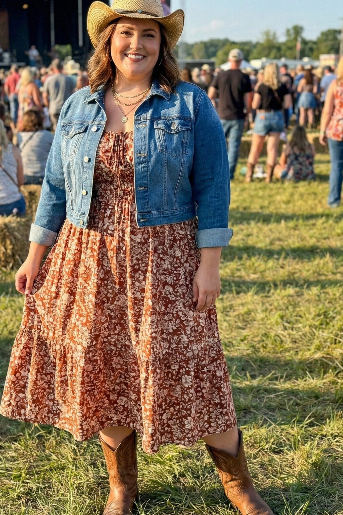 smiling woman at a festival in a straw cowboy hat, a medium-wash denim jacket over a rust floral midi dress and brown cowboy boots
