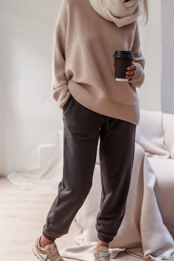 Beige sweater with dark joggers and sneakers, holding coffee - cozy casual outfit idea for women over 50