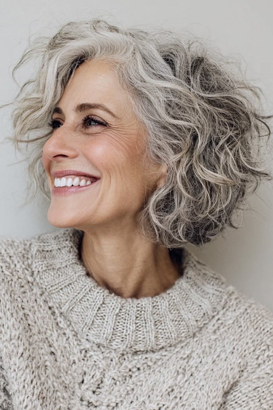 Silver curly bob on a laughing older woman in a cream sweater, side view, curly hairstyles for women over 50