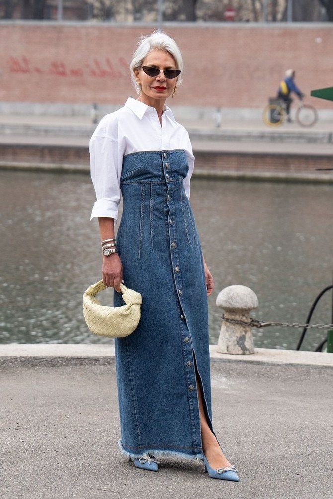 Older woman with white hair and sunglasses, white shirt layered under strapless button-front denim maxi dress, holding straw bag, standing by canal - denim dresses for women over 60