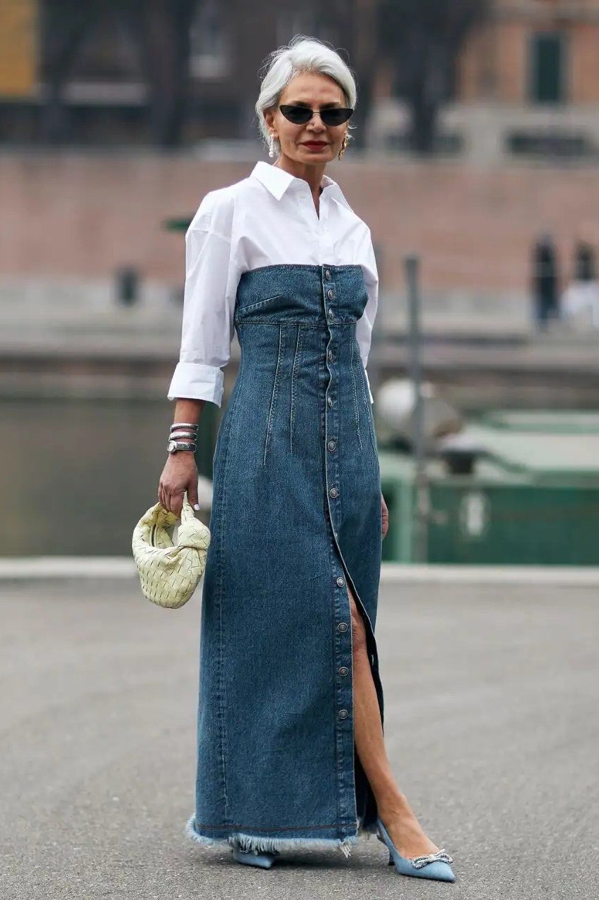 Same woman, strapless denim maxi dress with front slit over white shirt, walking pose with straw bag, city street - denim dresses for women over 60