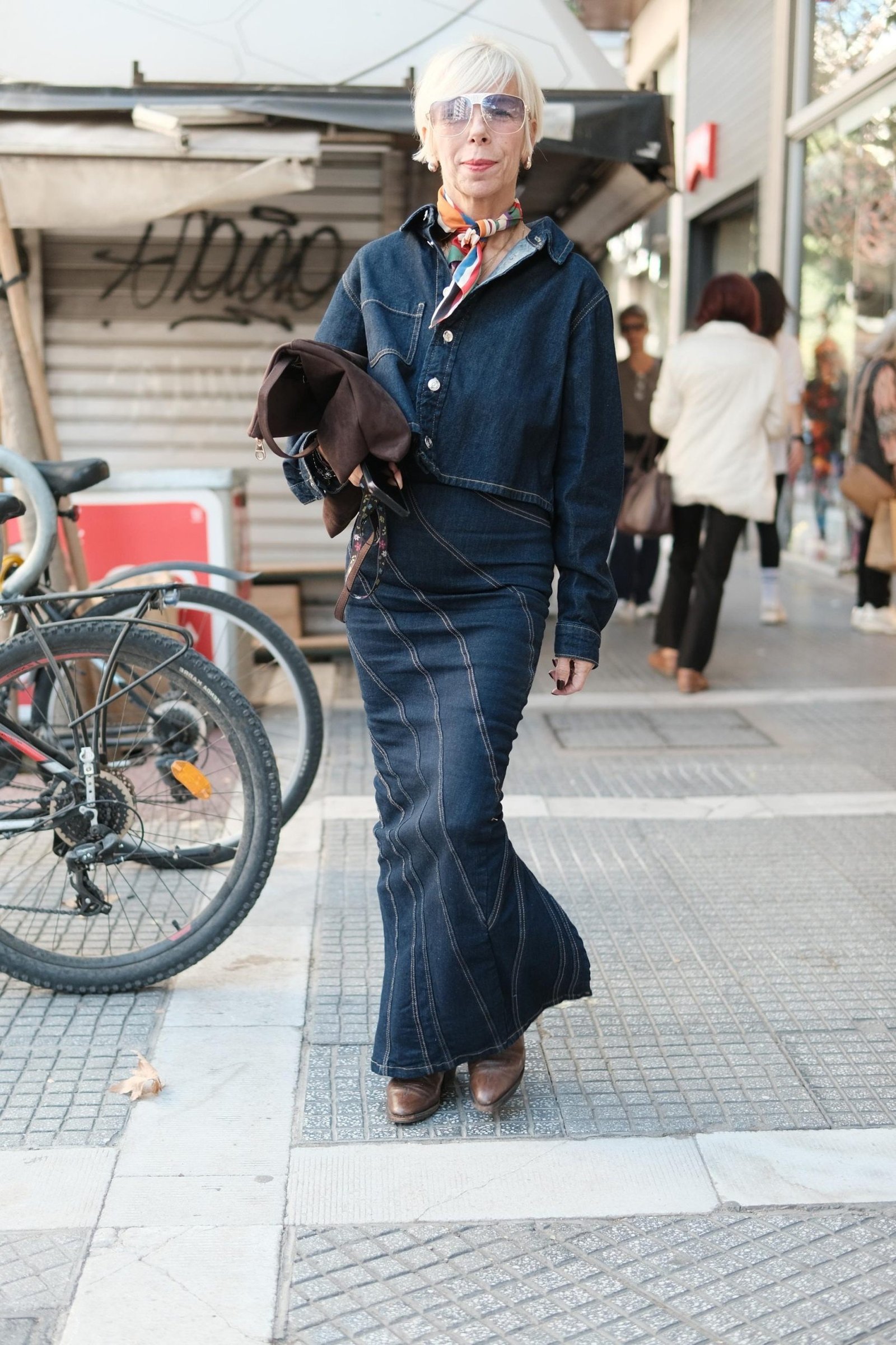 Older woman in dark denim shirt dress with sunglasses and dark bag, walking city street - denim dresses for women over 60