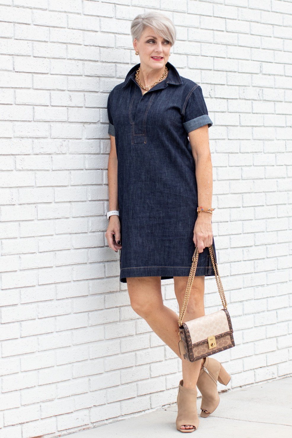 Older woman in dark denim short shirt dress with chain-strap handbag, posing against white brick wall - denim dresses for women over 60