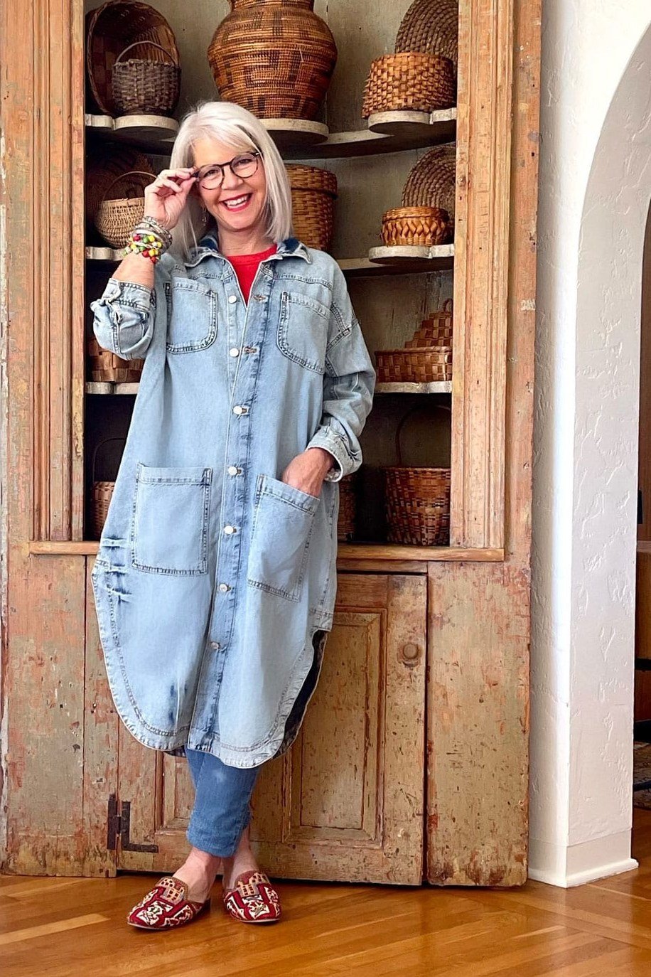 White-haired older woman in oversized light denim shirt dress with red sandals, by wooden hutch - denim dresses for women over 60