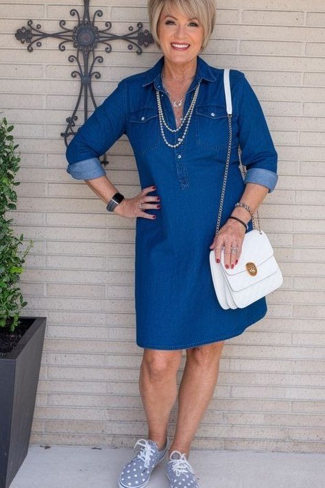 Blonde woman in dark denim shirt dress with white sneakers and white bag, by cross-decorated wall - denim dresses for women over 60