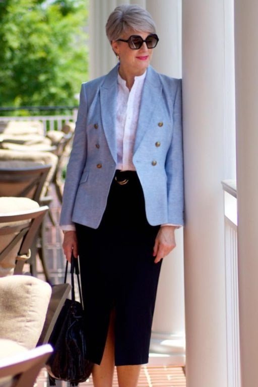 Elegant work outfit for women over 50: light blue blazer, white top, black midi skirt, sunglasses, leaning on column