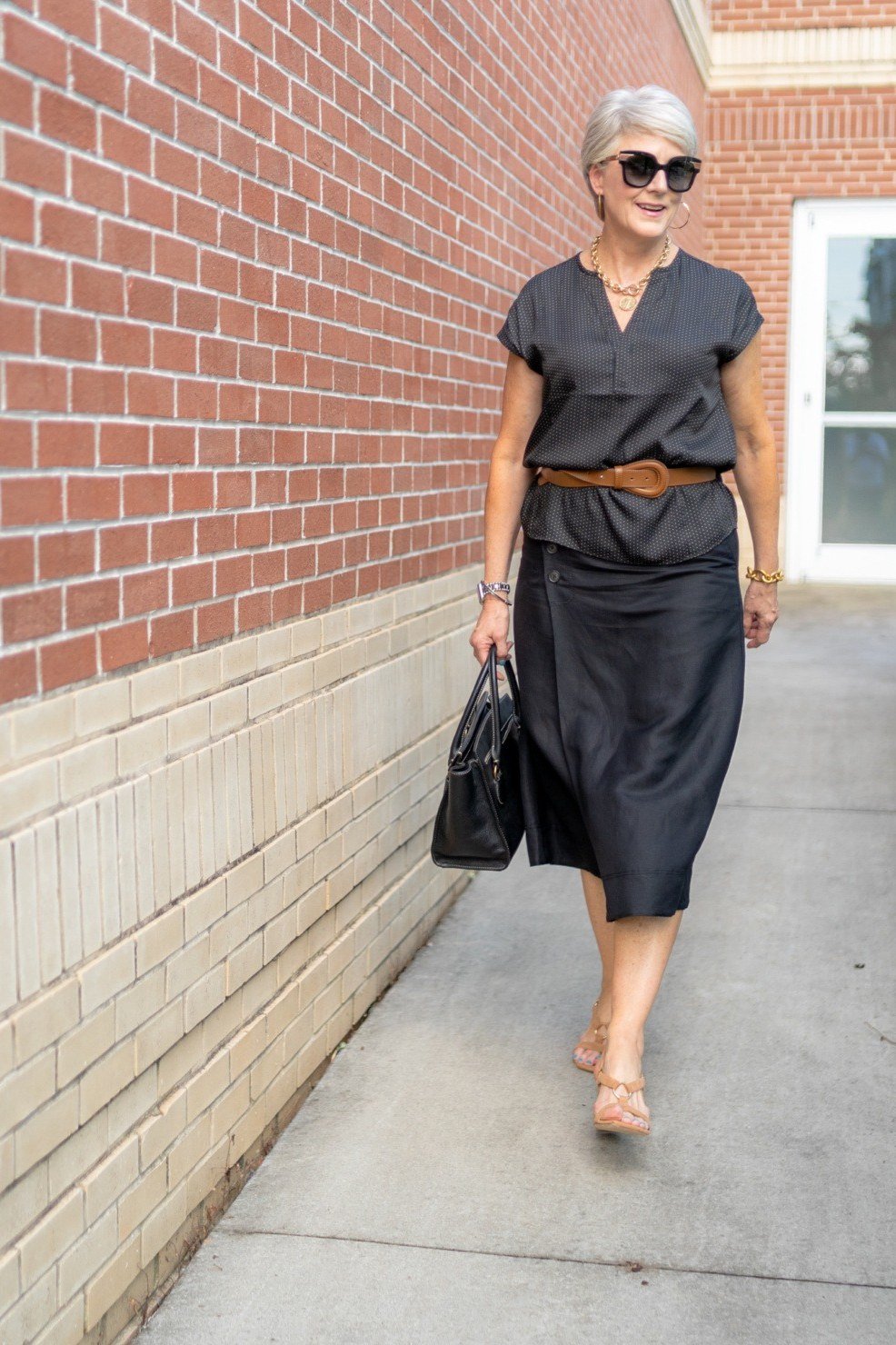 Elegant work outfit for women over 50: grey midi dress with belt, black bag, brick wall, full-body