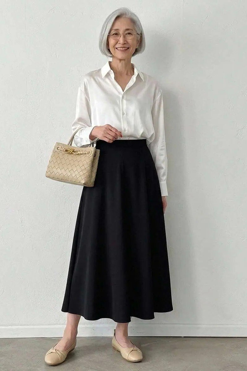 Elegant work outfit for women over 50: white blouse, black maxi skirt, woven bag, studio backdrop