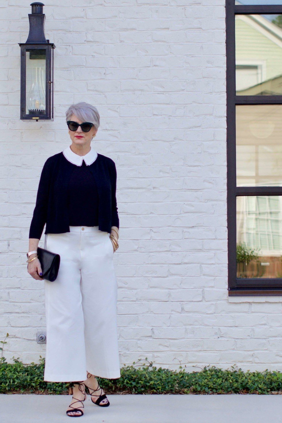 Elegant work outfit for women over 50: navy cardigan, white peter-pan collar top, white wide-leg culottes, sunglasses, black clutch, brick wall