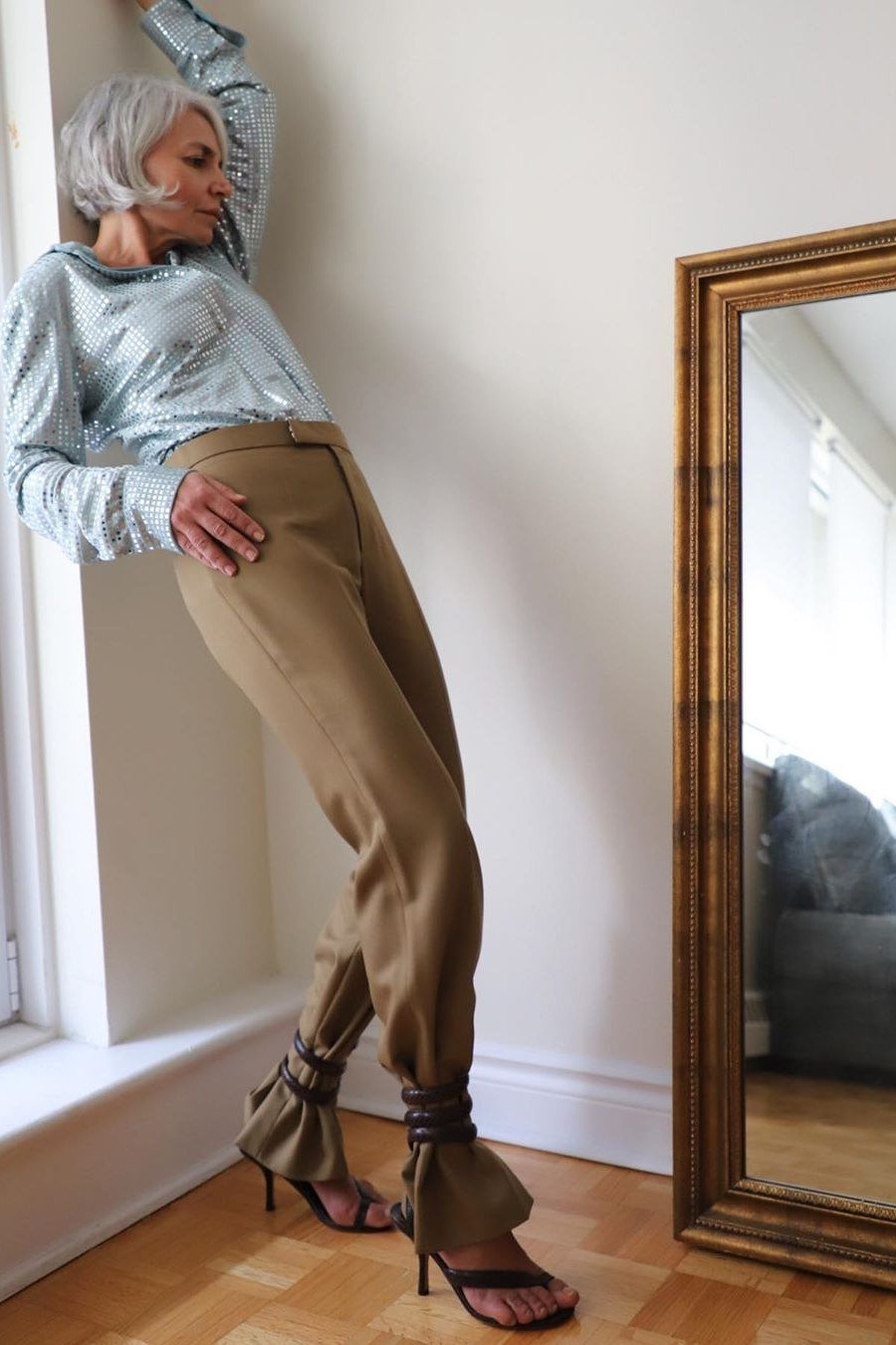 Elegant work outfit for women over 50: blue blouse, tan straight trousers, heels, leaning by window