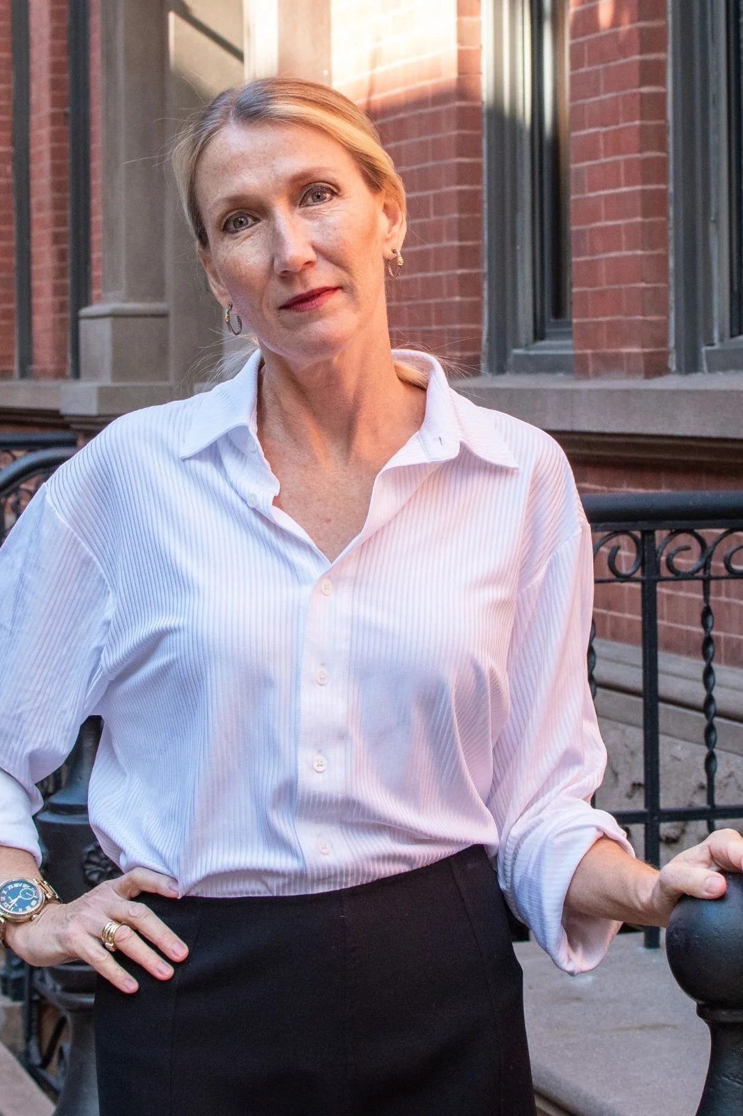 Elegant work outfit for women over 50: white shirt, black trousers, hand on hip, brick building, 3/4
