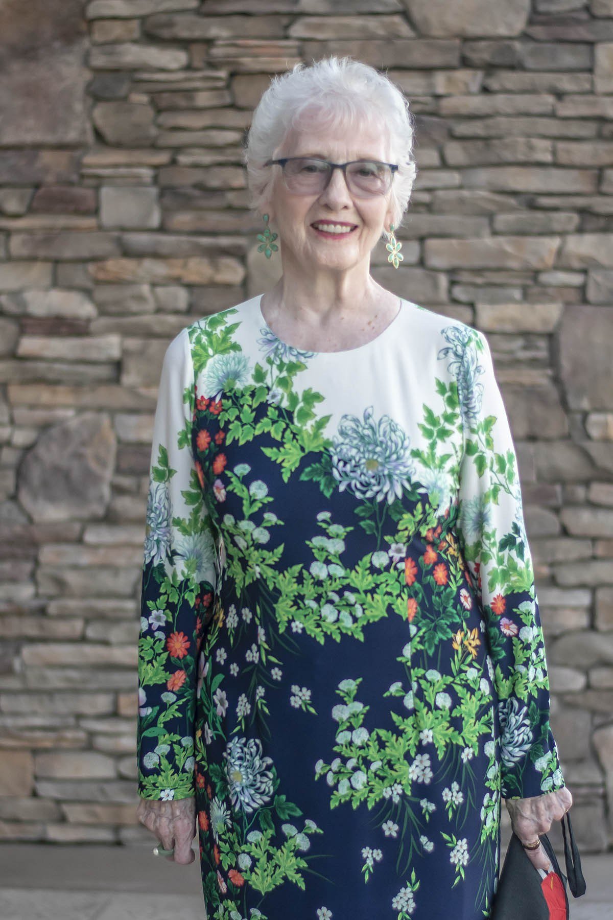 white floral blouse with dark floral midi skirt, glasses, older woman by stone wall - fall outfit idea for women over 70