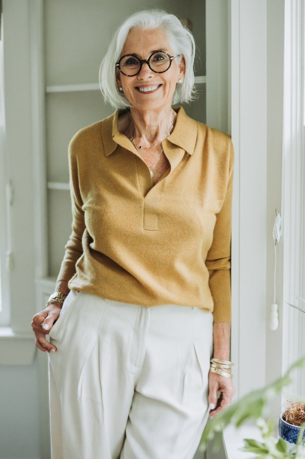 mustard polo sweater with white wide-leg trousers, older woman by window - fall outfit idea for women over 70