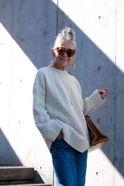 light gray sweater with blue jeans and sunglasses, older woman leaning on wall - fall outfit idea for women over 70