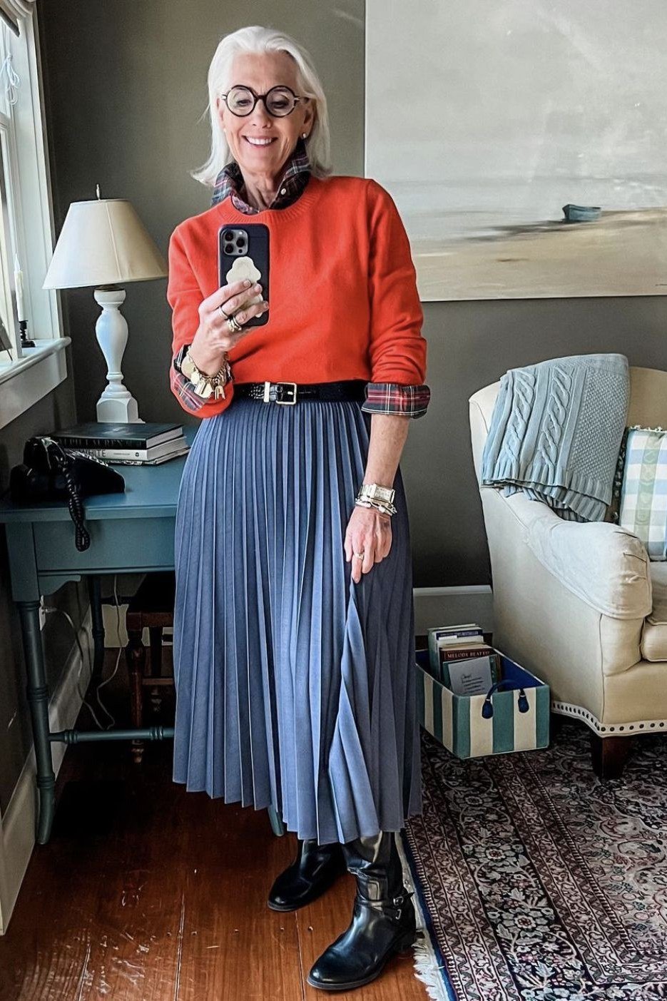rust orange sweater over plaid flannel shirt with blue pleated midi skirt and black boots, selfie indoors - fall outfit idea for women over 70