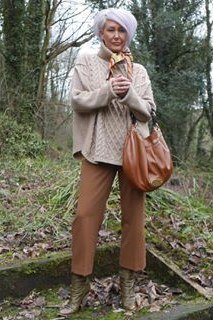 cream cable sweater with brown pants and tan bag in woods - fall outfit idea for women over 70