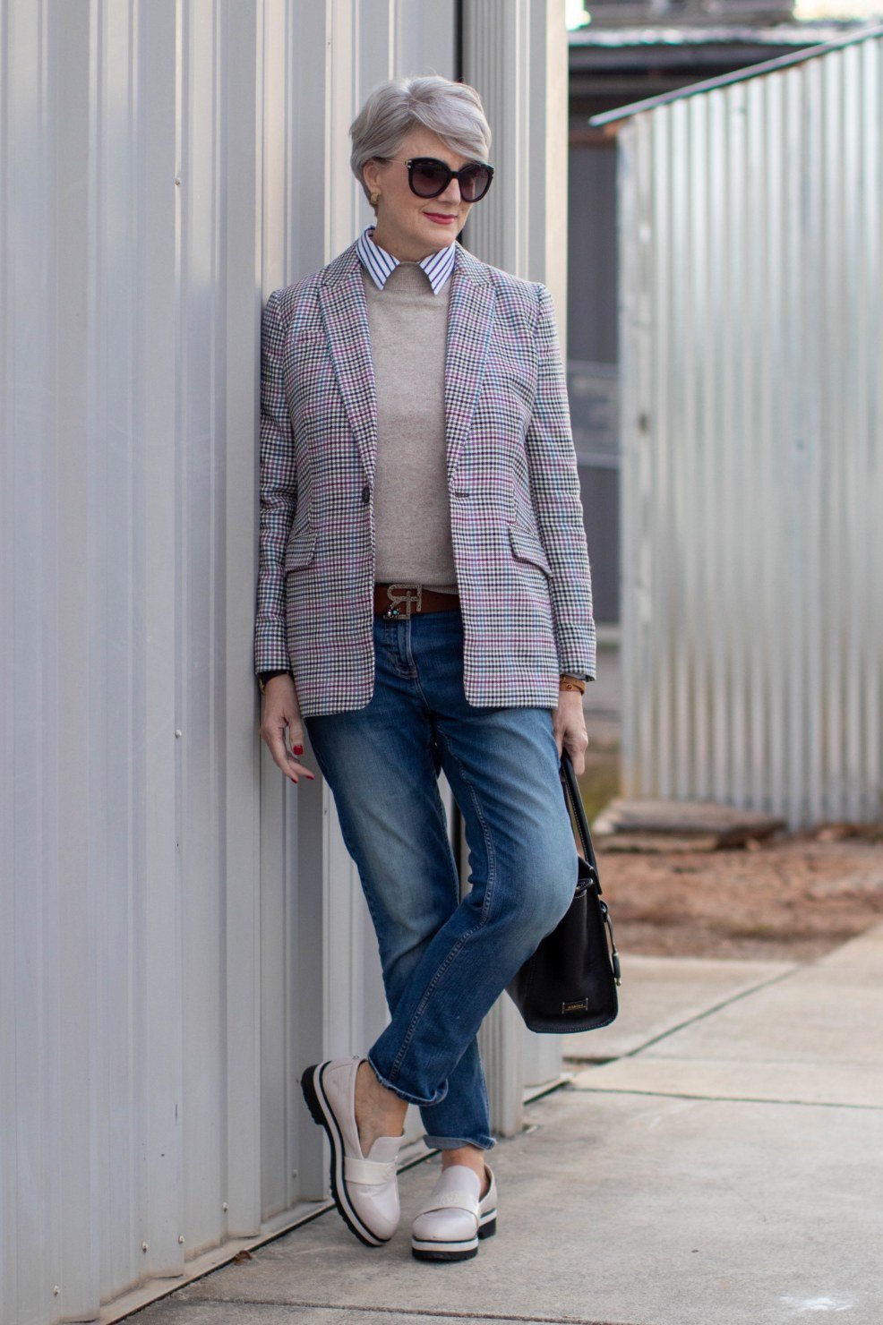 plaid blazer with gray top and blue jeans, older woman leaning on metal wall, white loafers and sunglasses - fall outfit idea for women over 70