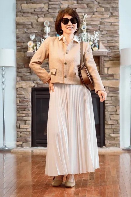 Camel cardigan over cream pleated midi skirt, fireplace, full body front view - fall cardigan outfit idea for women over 60