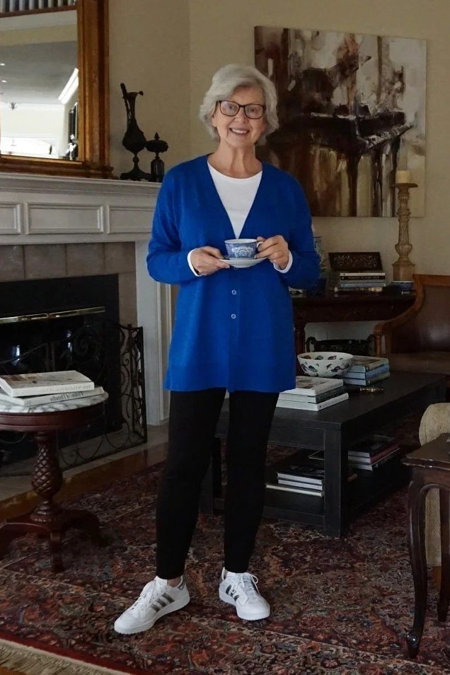 Royal blue cardigan over white top, black pants, holding cup, fireplace, full body front view - fall cardigan outfit idea for women over 60