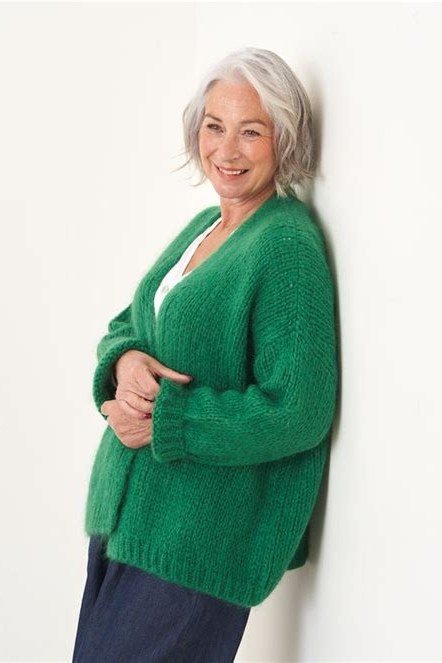 Emerald green chunky cardigan over white top, leaning against wall, 3/4 view - fall cardigan outfit idea for women over 60