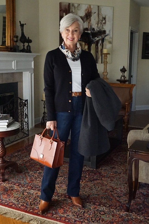 Black open cardigan over white top, blue jeans, brown ankle boots and tan tote, full body front view by fireplace - fall cardigan outfit idea for women over 60