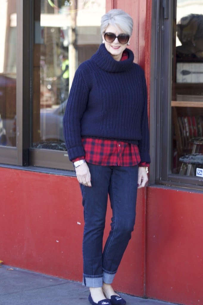 Woman over 60 wearing a navy cable sweater over a red plaid shirt with jeans