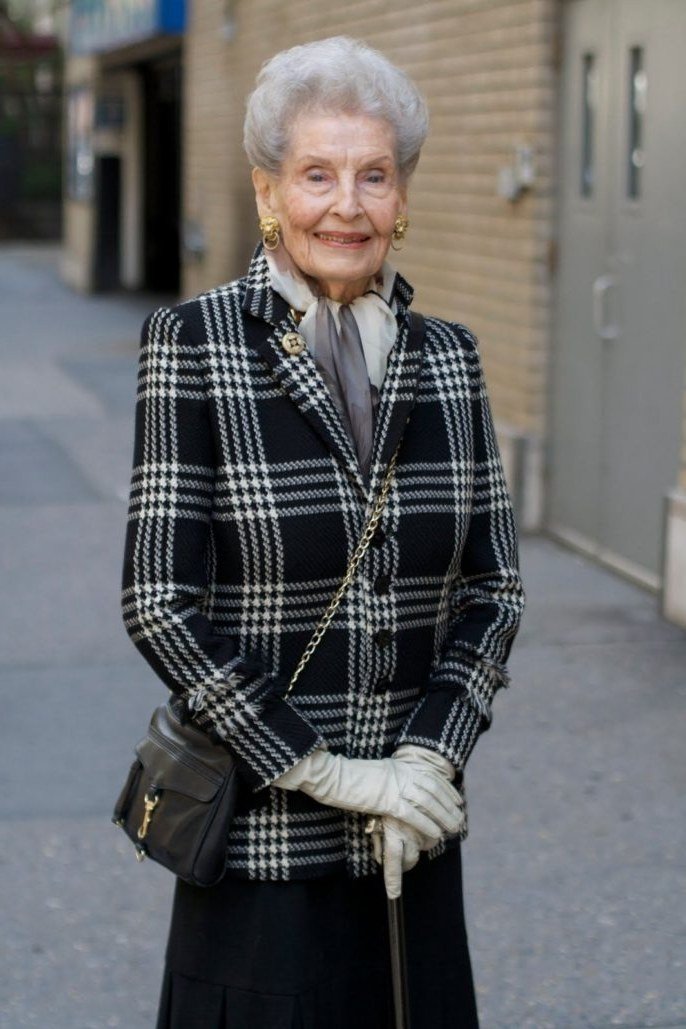 Older woman in a black and white plaid check jacket with a gray scarf, gloves and crossbody bag