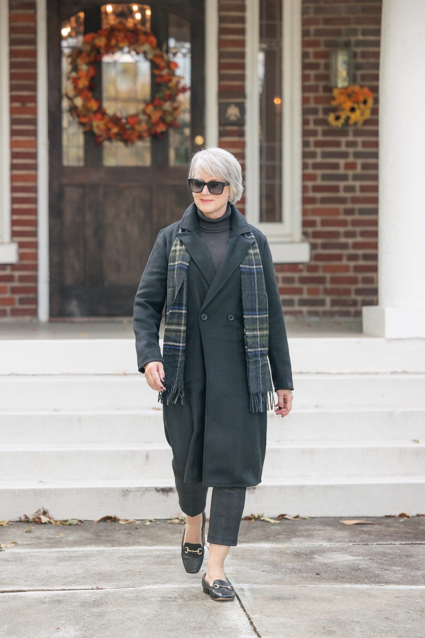 Older woman with gray hair wearing a dark plaid coat and tartan scarf with sunglasses on a white doorstep - stylish fall look for older women