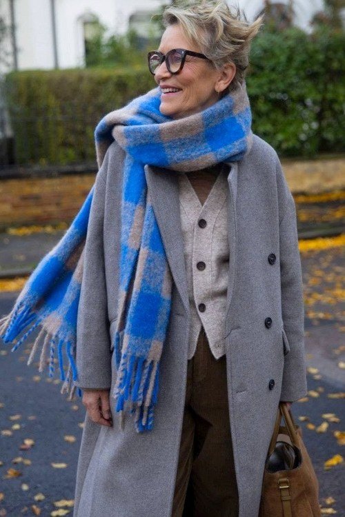 Older woman with short gray hair wearing a gray coat and bright blue plaid scarf on an autumn street - stylish fall look for older women