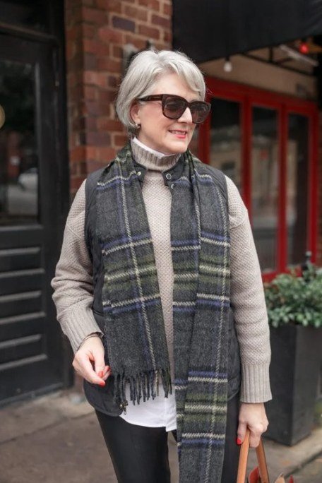 Older woman in a beige turtleneck with a long green plaid vest and sunglasses by a brick storefront - stylish fall look for older women