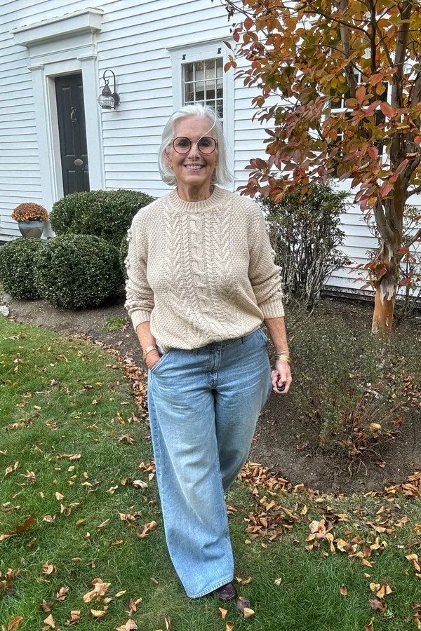 Older woman in a cream cable-knit sweater with wide-leg jeans on a lawn - stylish fall look for older women