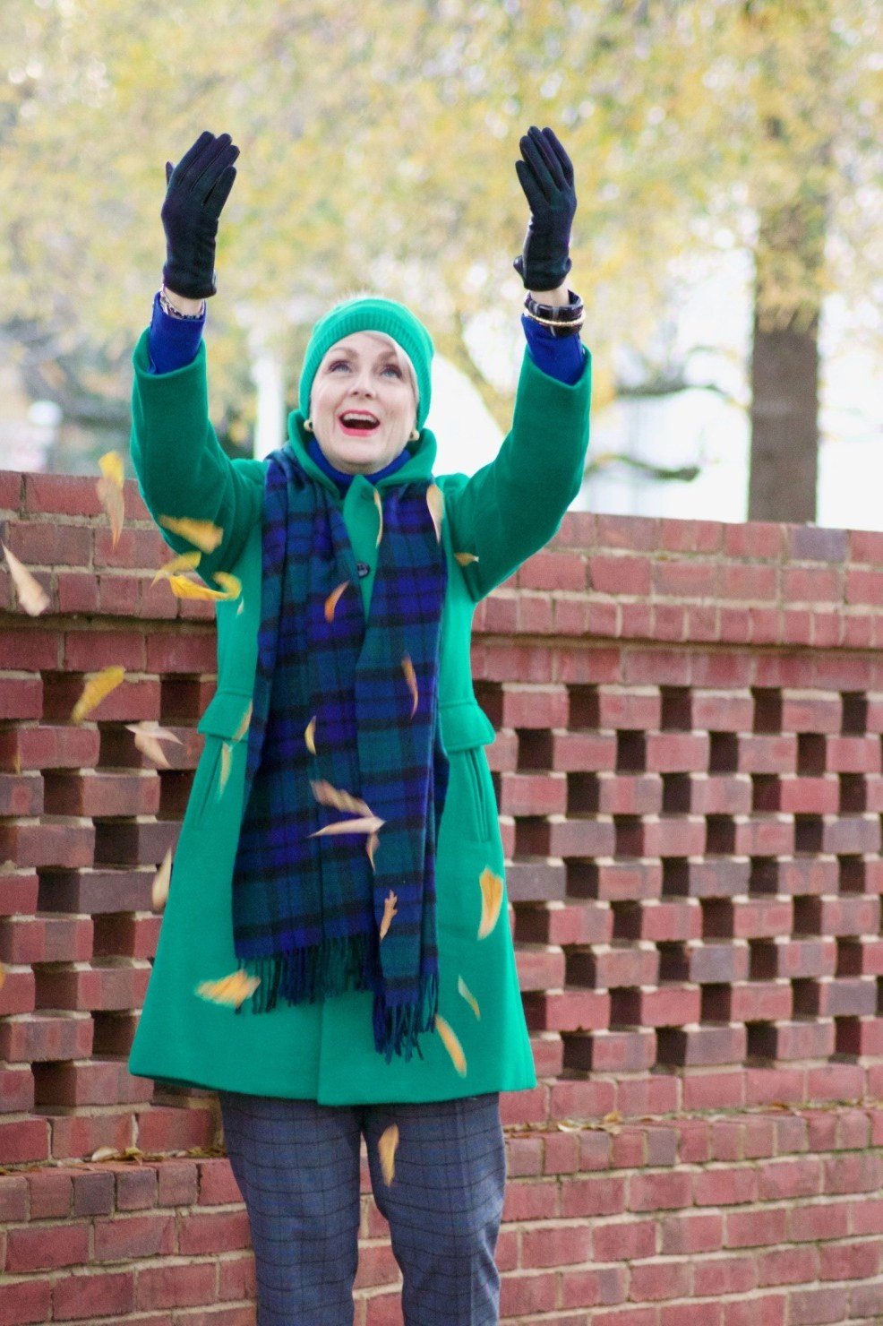 Older woman in an emerald green coat with a blue plaid scarf, arms raised playfully against a brick wall - stylish fall look for older women