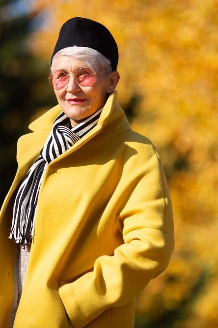 Older woman in a mustard yellow coat with a striped scarf and black cap in an autumn park - stylish fall look for older women