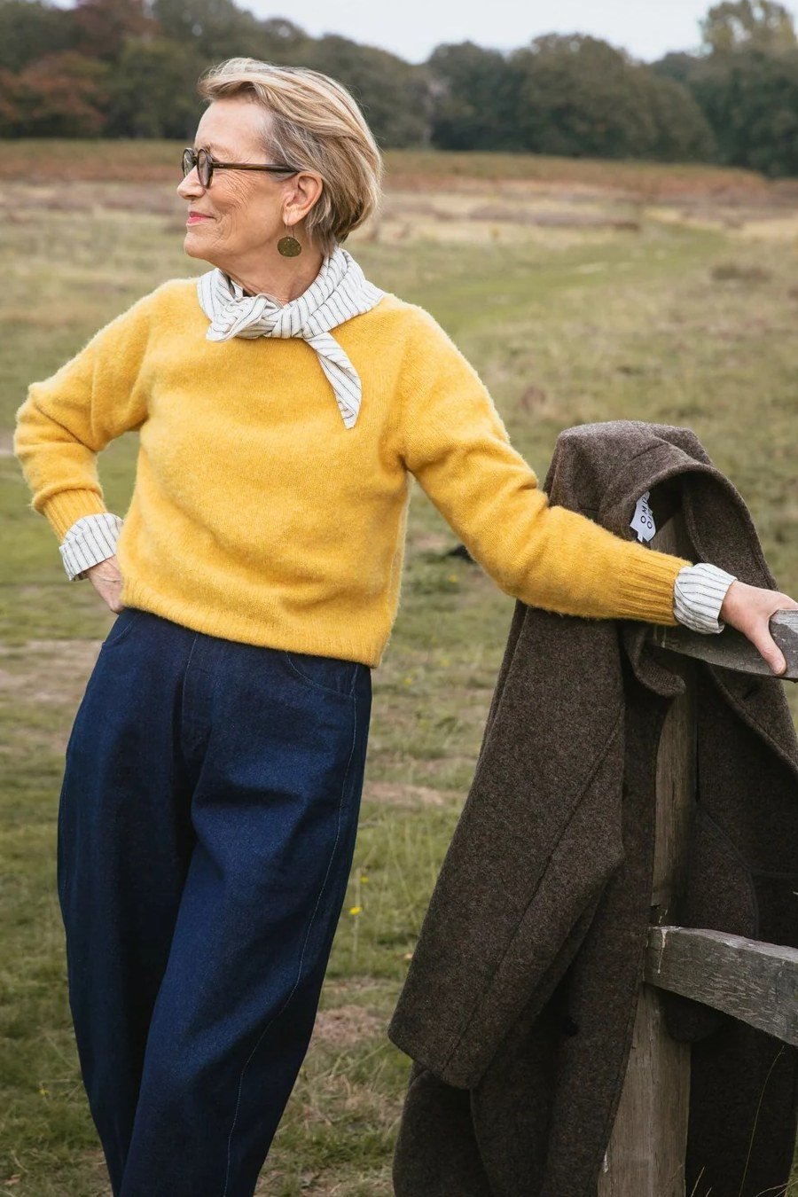Older woman in a mustard sweater with navy wide-leg trousers, holding a coat, in a field - stylish fall look for older women