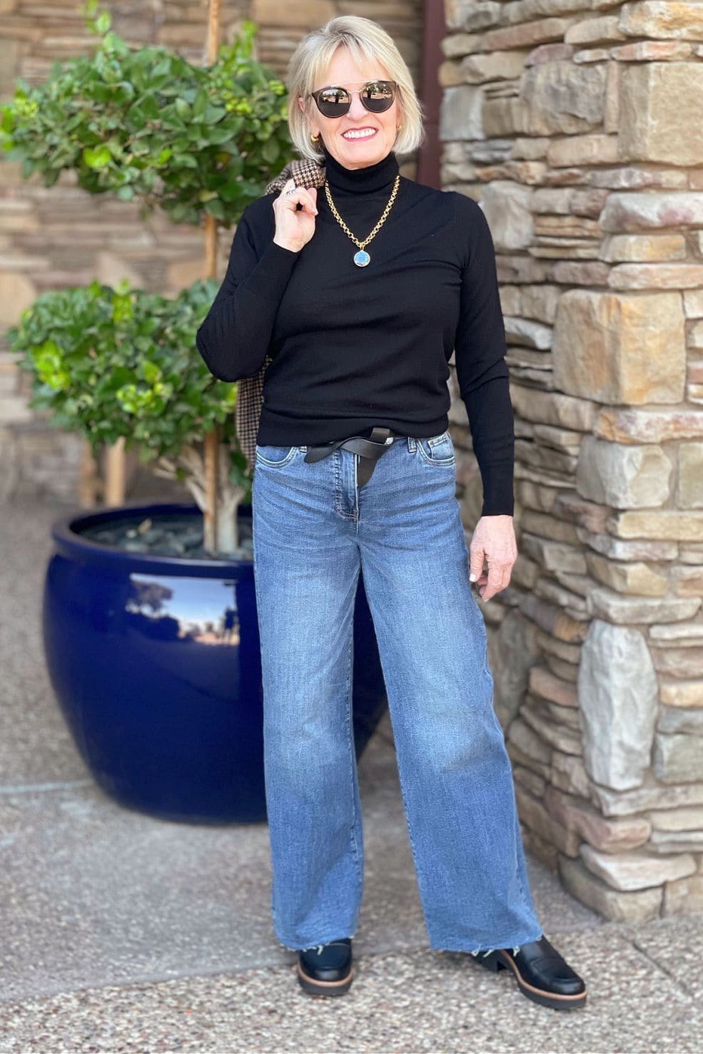 A fitted black turtleneck tucked into light-wash wide-leg jeans makes a clean, modern line, standing by a stone wall beside a blue planter. The dark top against pale denim gives a slimming color-block effect. Sunglasses and a simple necklace finish it. This works for lunch, errands, or a gallery visit.