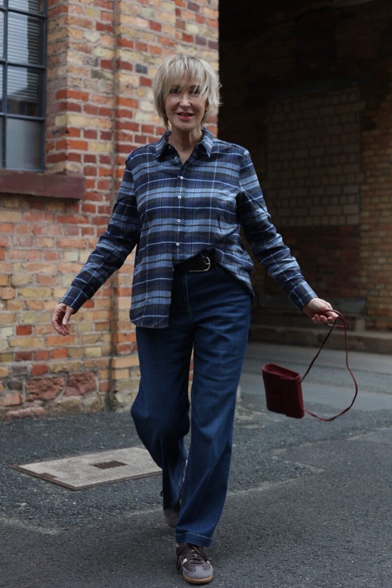 A blue plaid shirt with dark jeans gets dressed up with a small burgundy handbag. In a brick alley, the colors feel rich and autumnal. Short gray hair looks striking with blue tones. A polished casual look for city days.