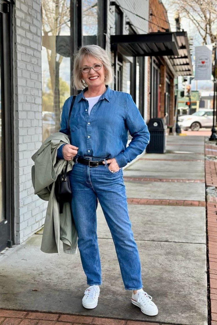 A chambray shirt with medium jeans and white sneakers is fresh and sporty. An olive jacket draped over one arm adds a layer for cool sidewalks. Sunglasses complete the easy vibe. A great travel or sightseeing outfit.