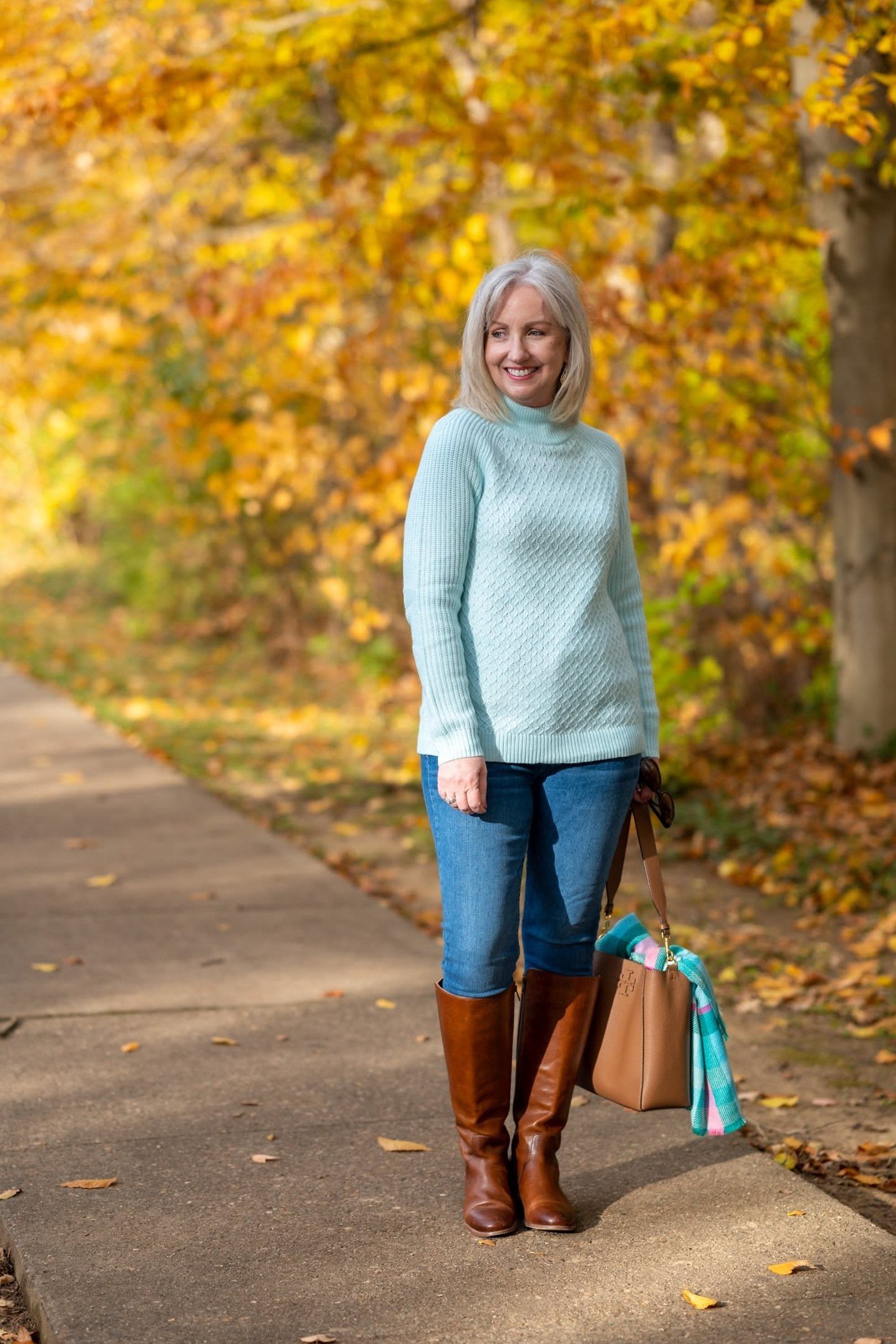 A light blue turtleneck with medium jeans, brown knee boots, and a tan bag is fresh fall color. The pale blue brightens an autumn day. The contrast against fall foliage is pretty. A cheerful outfit for daytime outings.
