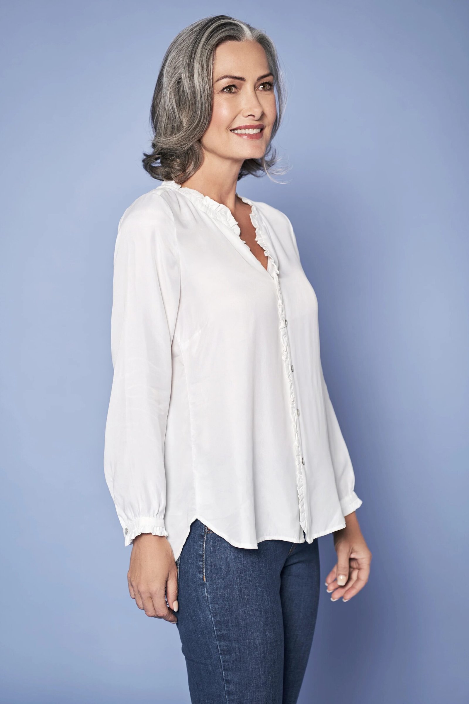 A white blouse with medium jeans, photographed against a light blue background, is fresh and pretty. The blouse has a soft, feminine drape. Gray hair looks radiant with crisp white. A beautiful daytime look for luncheons.