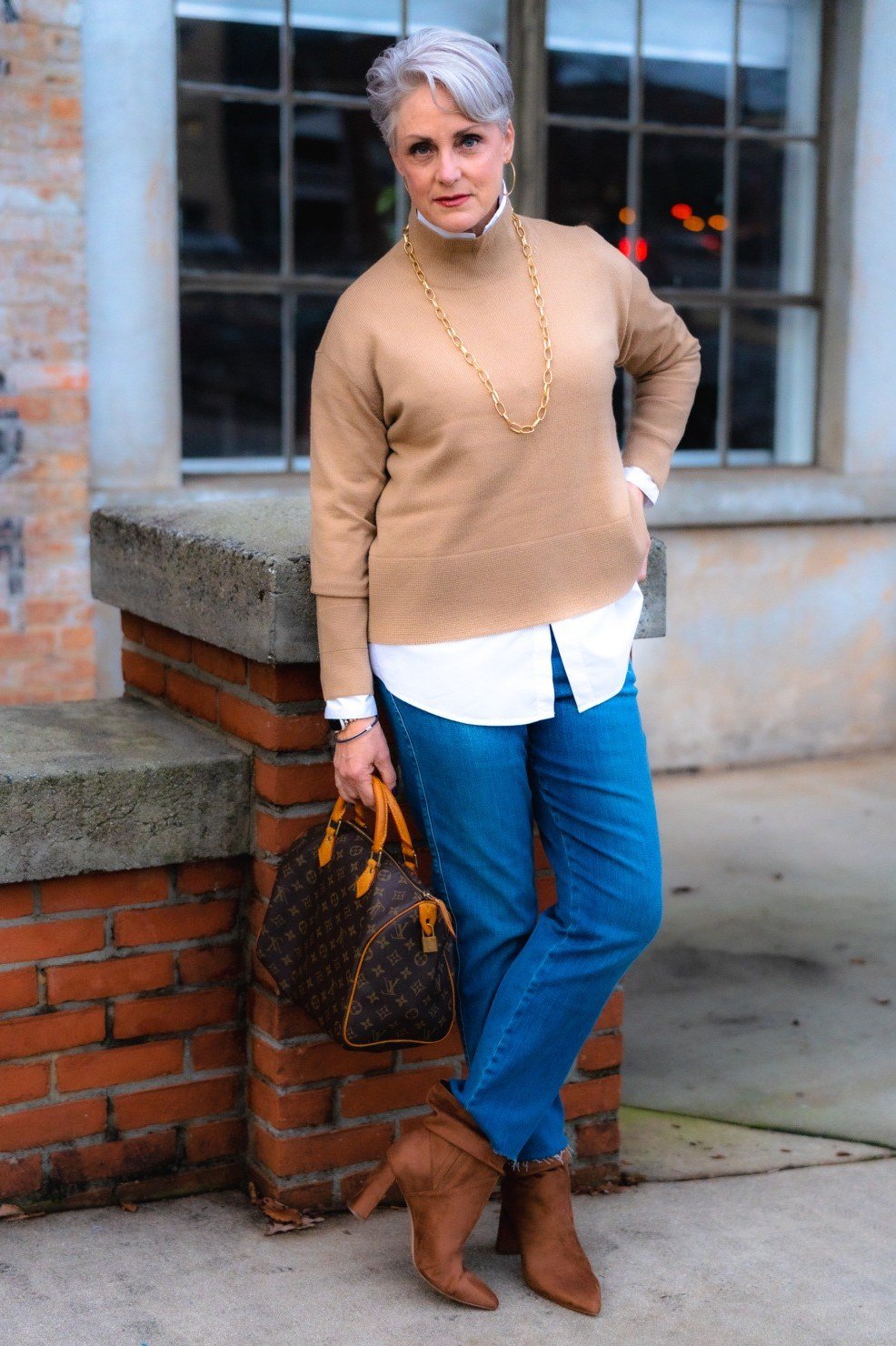 Camel sweater over white shirt, blue jeans, tan ankle boots, older woman at brick wall