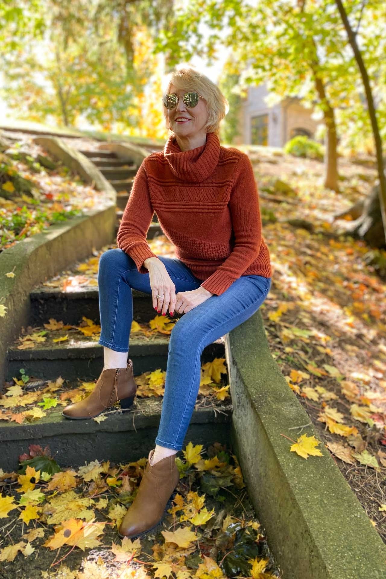 Rust turtleneck sweater, blue jeans, tan ankle boots, woman on leafy stone steps