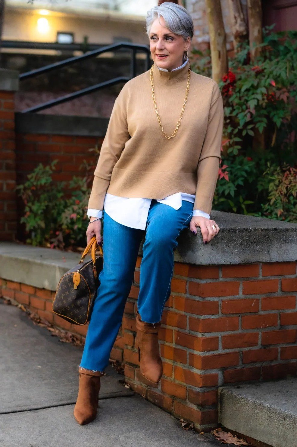 Tan sweater over white shirt, blue jeans, tan boots, woman at brick wall