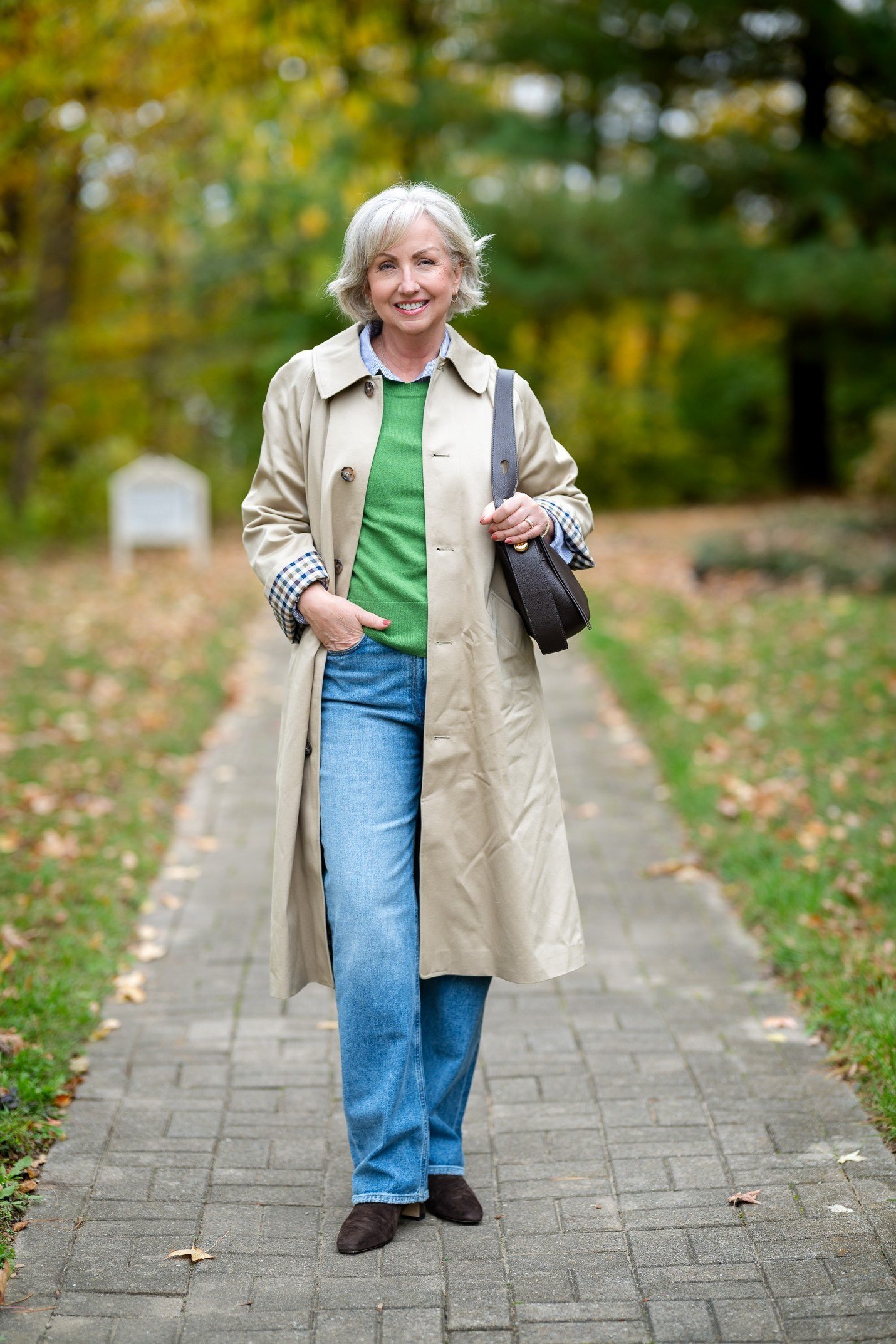 Beige trench coat, green sweater, blue jeans, woman on brick path