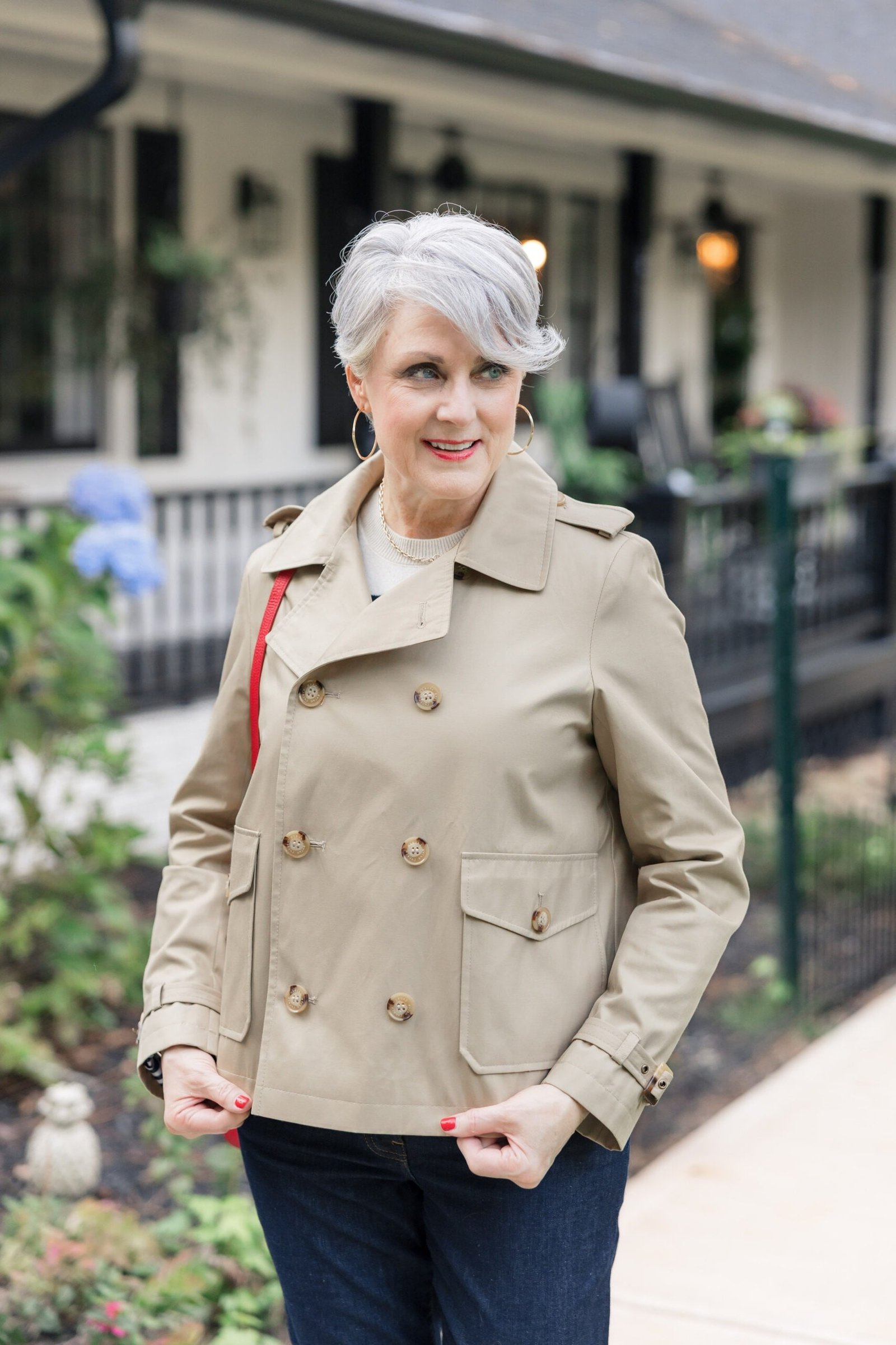 Classic beige trench coat, white top, dark jeans, woman on porch